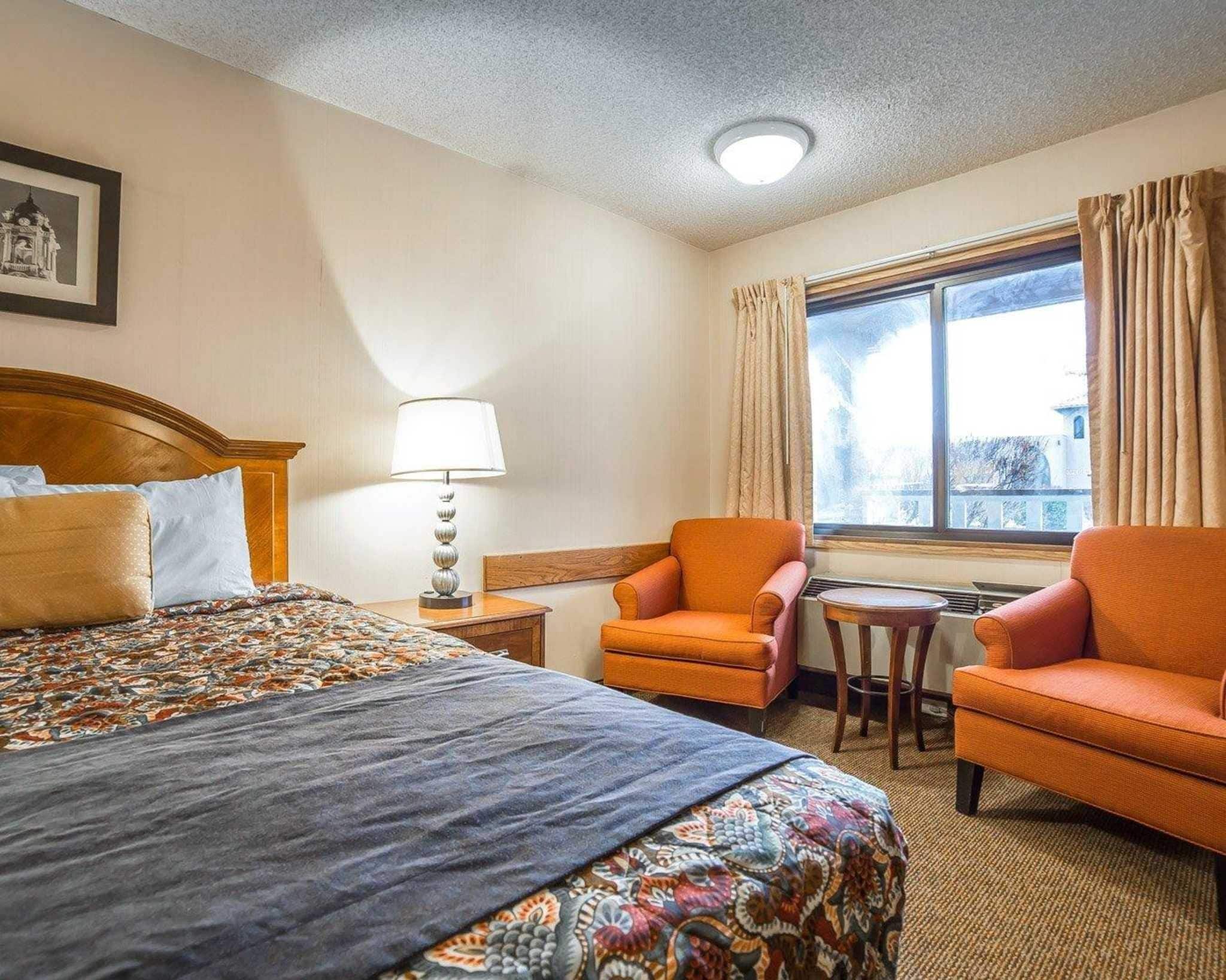 A hotel room with a bed, two armchairs, a small table, a lamp, and a window with curtains.