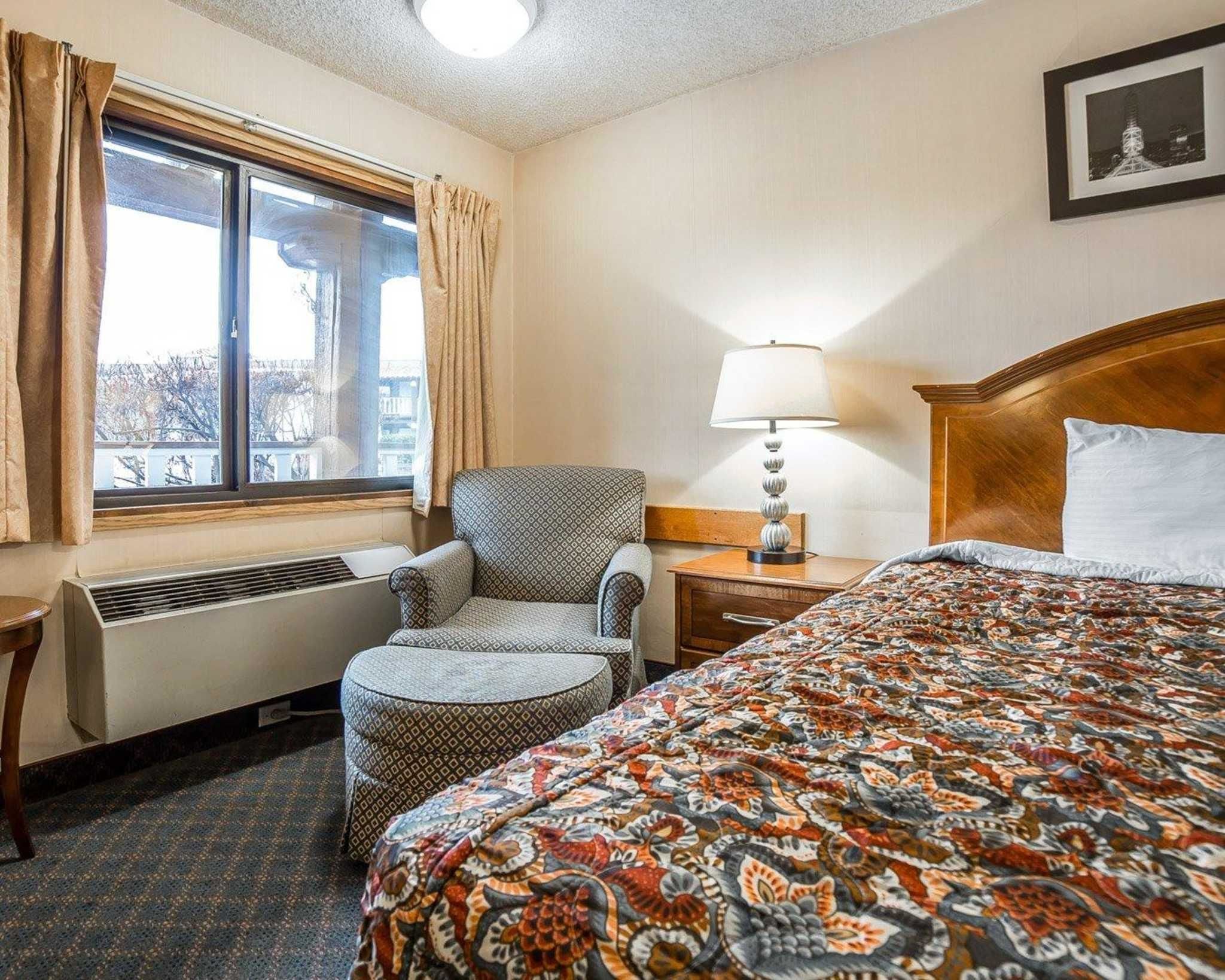 Une chambre d’hôtel comprenant un lit, une chaise, une table de chevet avec une lampe, une fenêtre avec des rideaux et un radiateur.