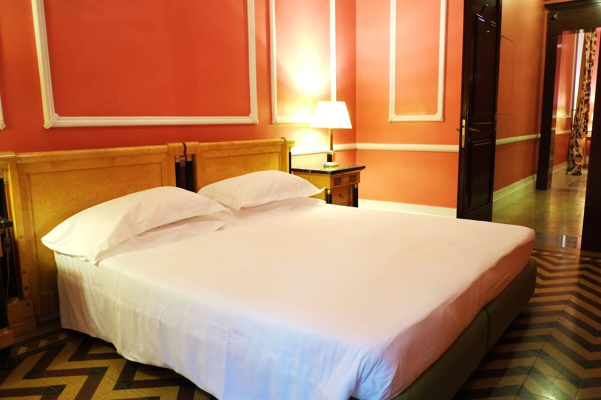 Hotellihuone, jossa on suuri sänky, yöpöytä lampun kanssa ja ovi, joka johtaa toiseen huoneeseen.