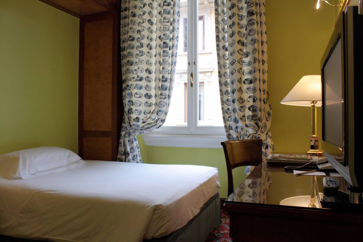 Hotellihuone, jossa on sänky, tuoli, kirjoituspöytä lampun kanssa ja televisio.