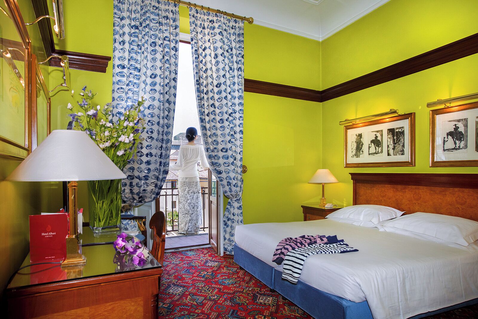 Habitación de hotel con cama, dos lámparas de noche, un libro rojo sobre un escritorio, un jarrón con flores y vistas a través de una ventana con cortinas azules.