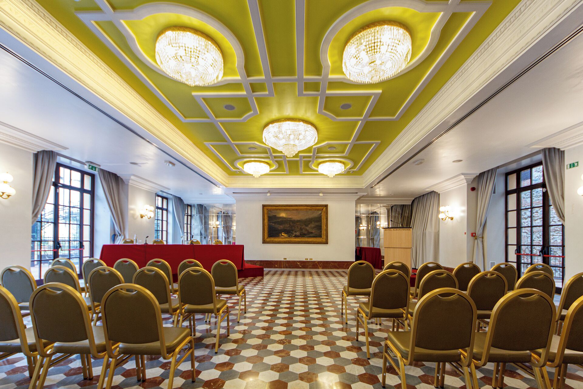 A conference room with a checkered floor, rows of chairs, a red podium, and chandeliers.