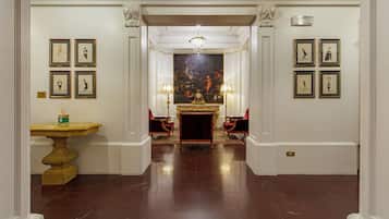 A grand hallway with marble floors, framed artwork on the walls, a central painting, and a chandelier.