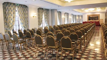 Un salón de conferencias con filas de sillas y mesas, ventanas amplias con cortinas, y un suelo con diseño.