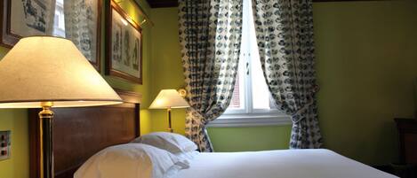 Habitación de hotel con cama, dos lámparas de noche, teléfono y ventana con cortinas estampadas.