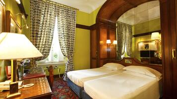 A hotel room with two beds, a desk with a lamp, a telephone, and a window with curtains.