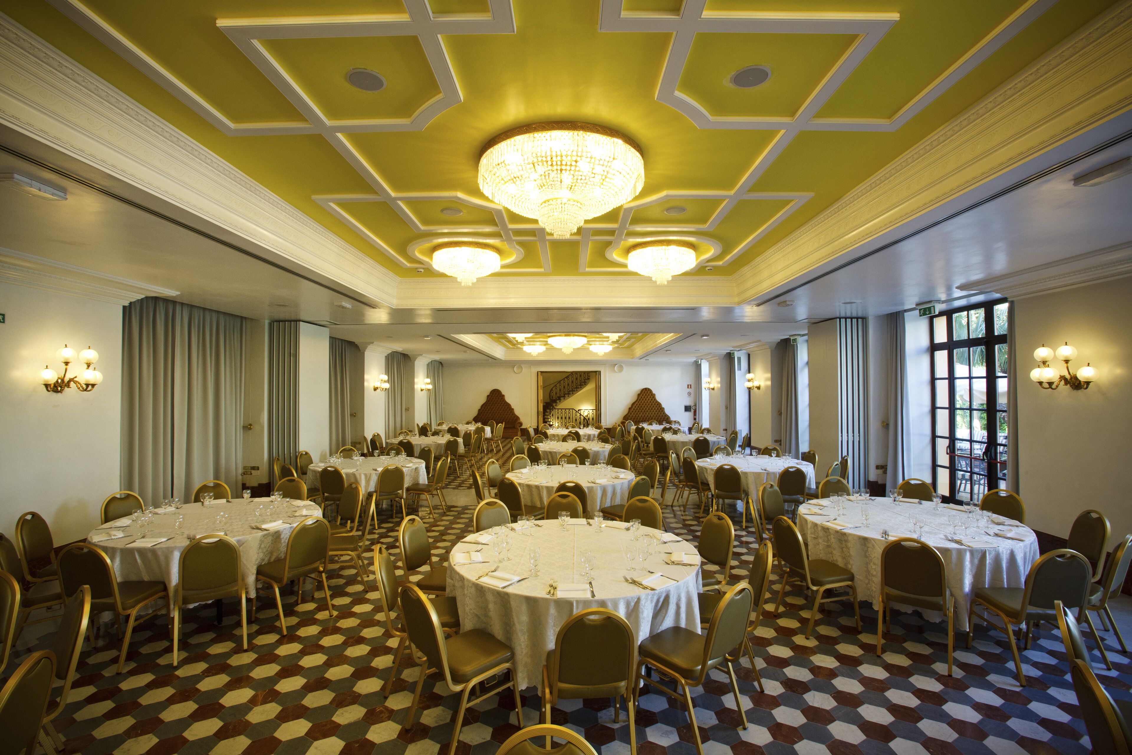 Un amplio salón de banquetes con mesas redondas, sillas doradas y una lámpara de araña.