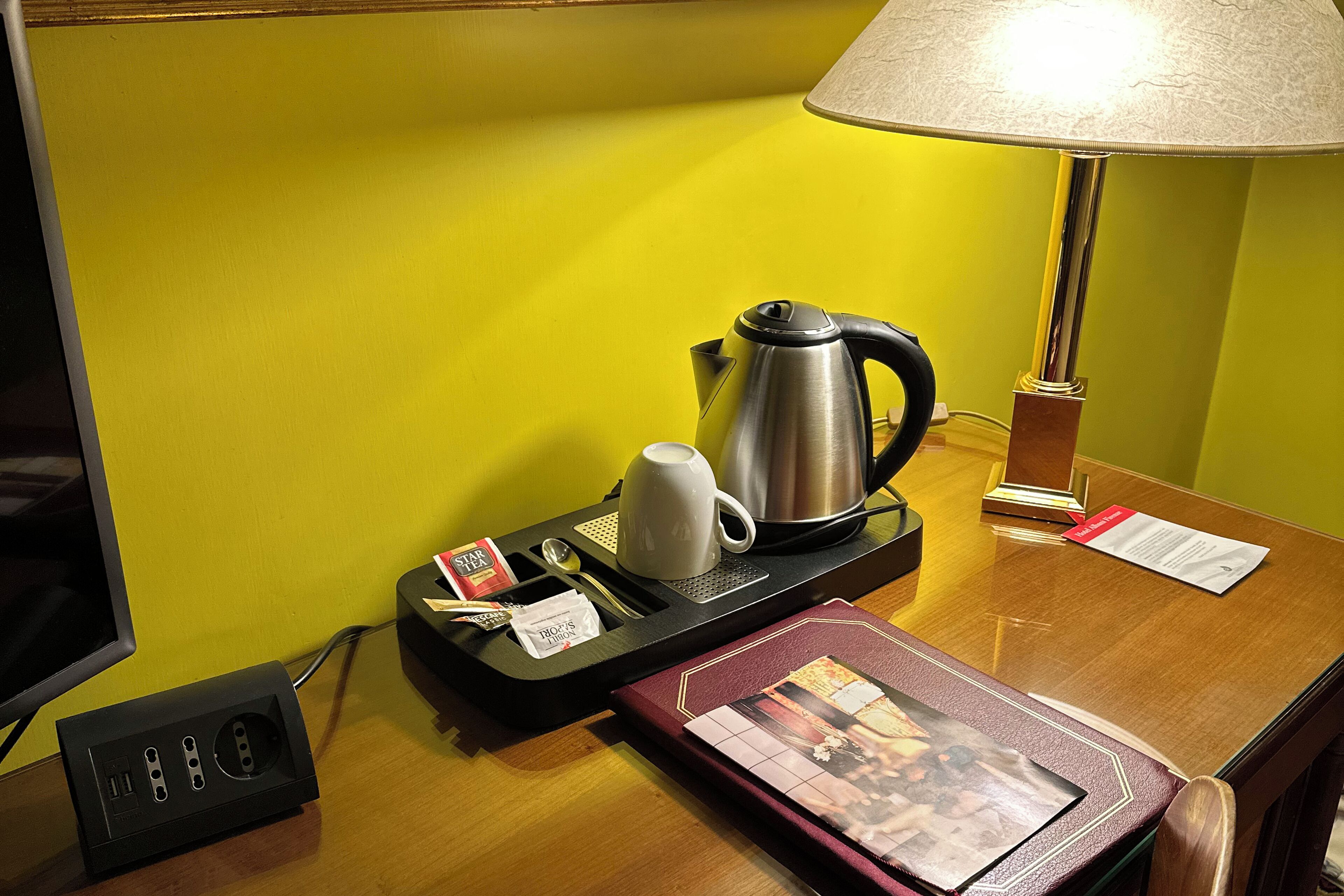 A hotel room with a wooden desk, a lamp, a kettle, a mug, a tray with tea bags, a book, and a TV.