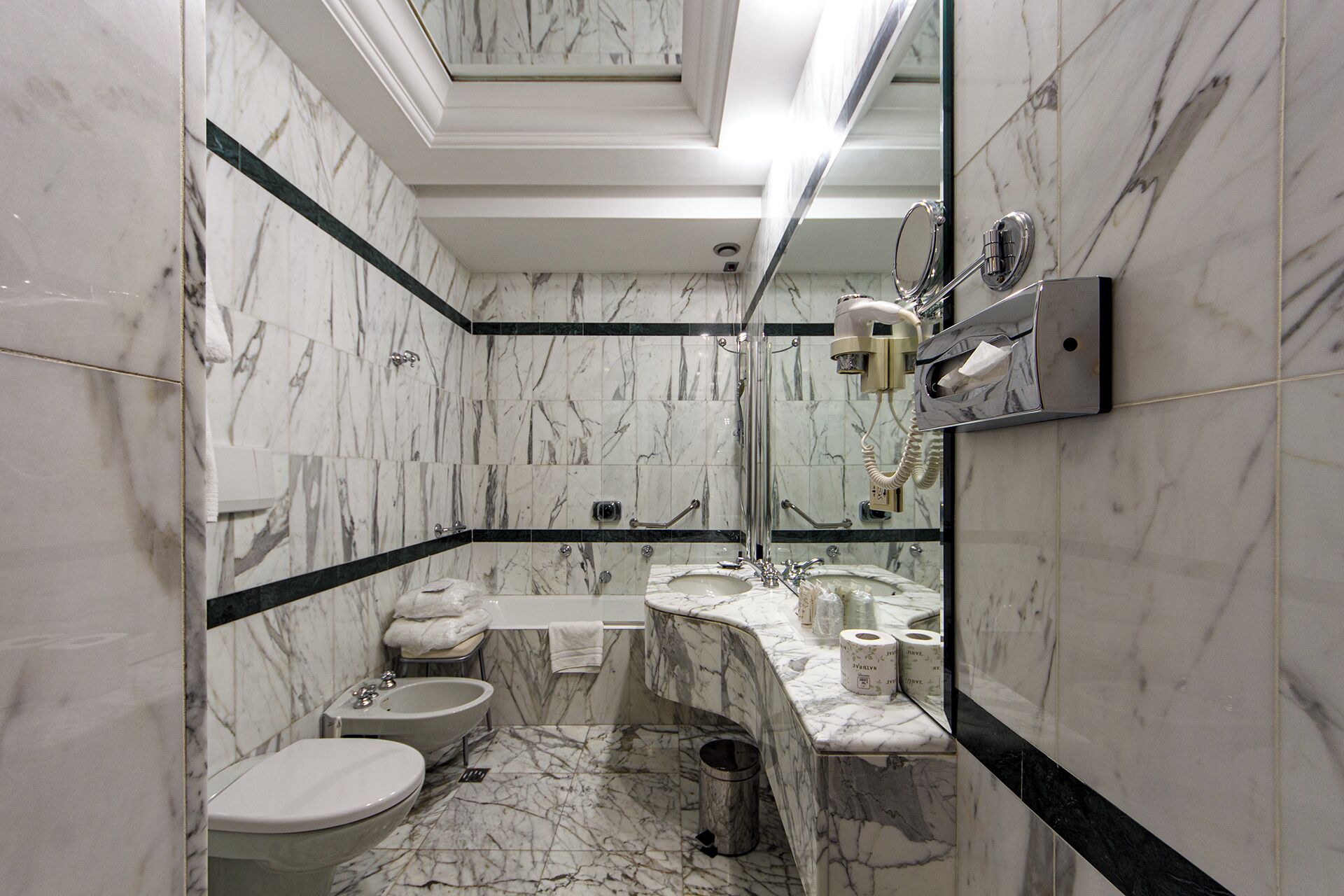 A bathroom with a marble floor, a toilet, a bidet, a sink, a mirror, and a hairdryer mounted on the wall.