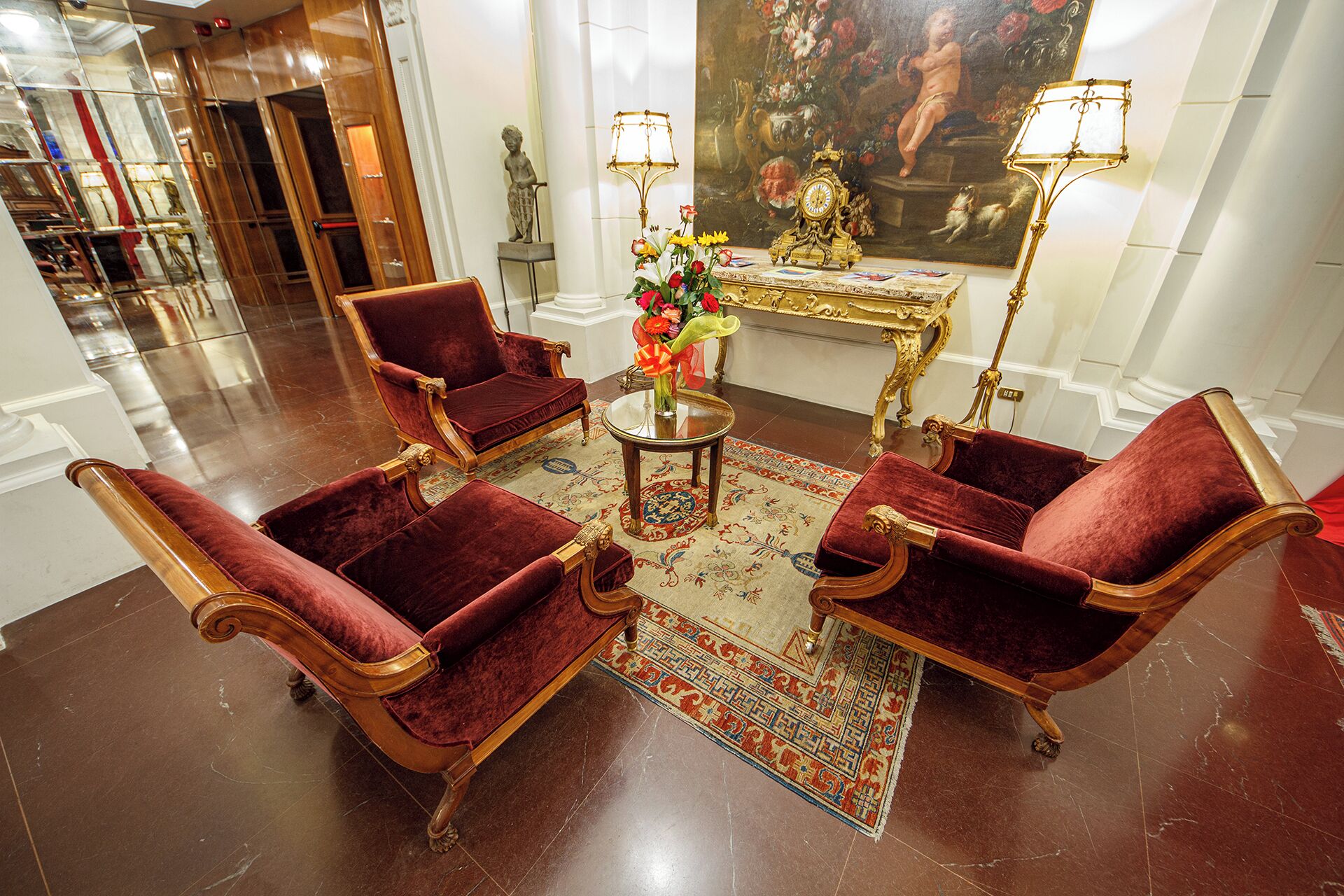 A lobby with four red velvet armchairs, a small round table with a vase of flowers, and two ornate lamps.