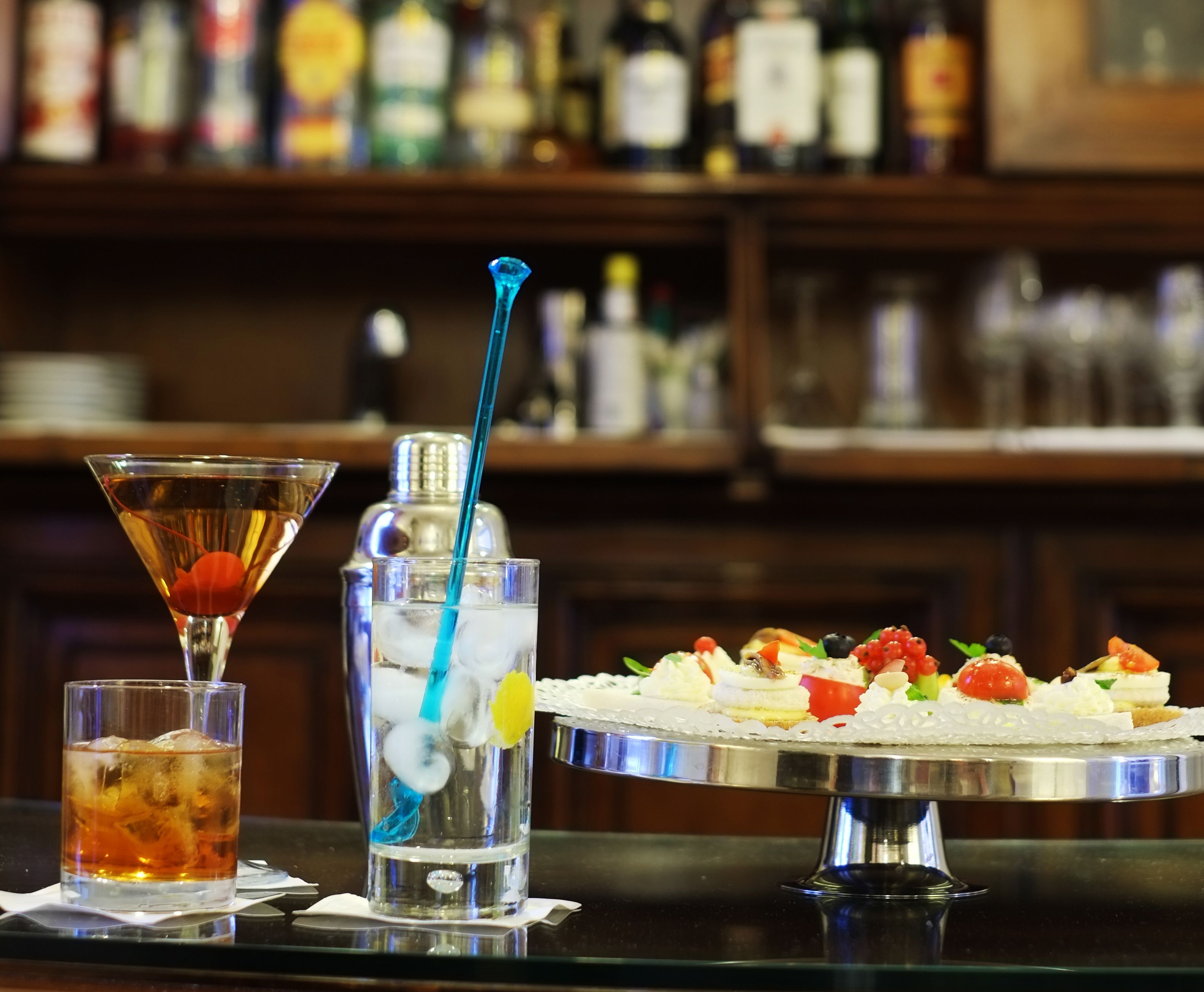 Una zona de bar con una copa de martini, una jarra con un popote azul y una bandeja de aperitivos.