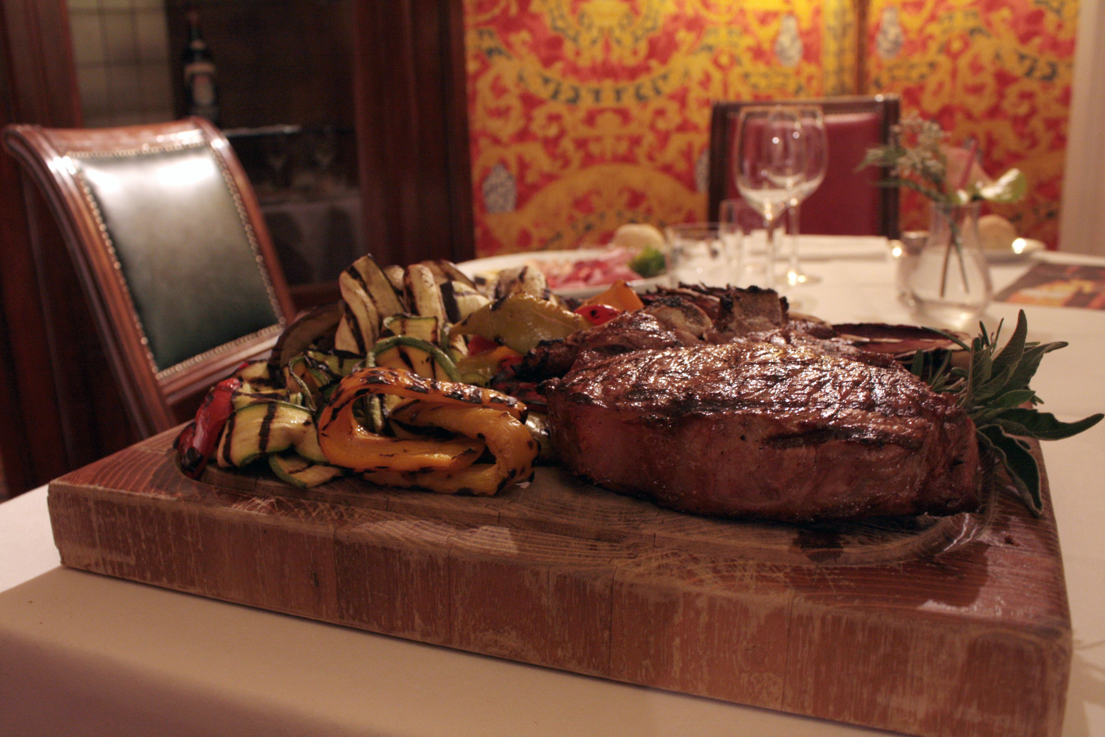 A steak with vegetables on a wooden board, a glass of wine, and a vase with flowers on a table.