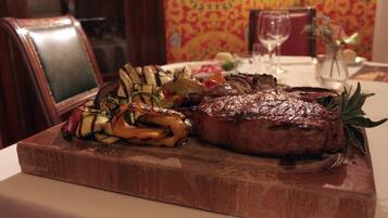 A steak with vegetables on a wooden board, a glass of wine, and a vase with flowers on a table.