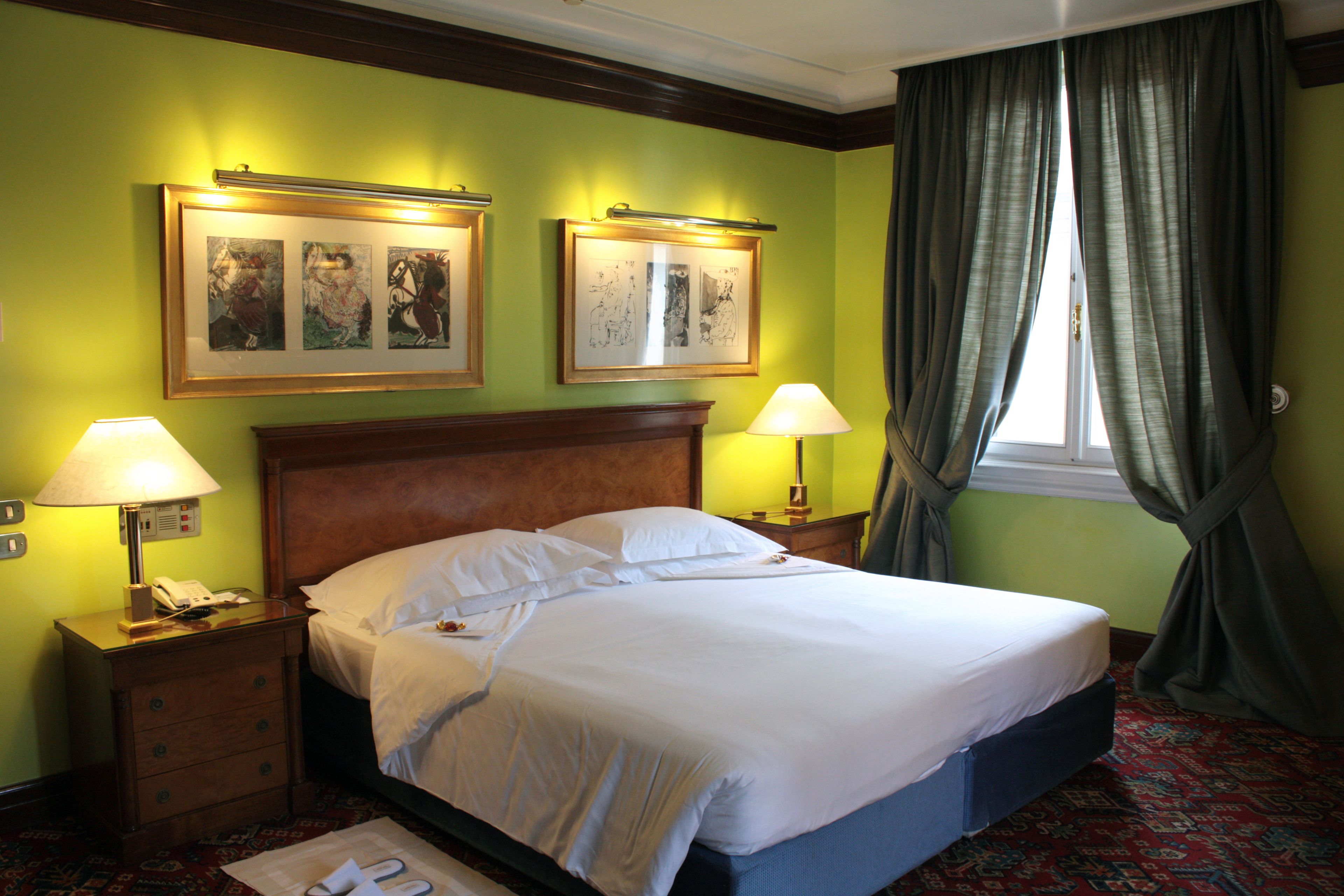 Un letto rifatto con cura in una camera d'albergo con pareti verdi, due quadri appesi sopra e una finestra con tende.