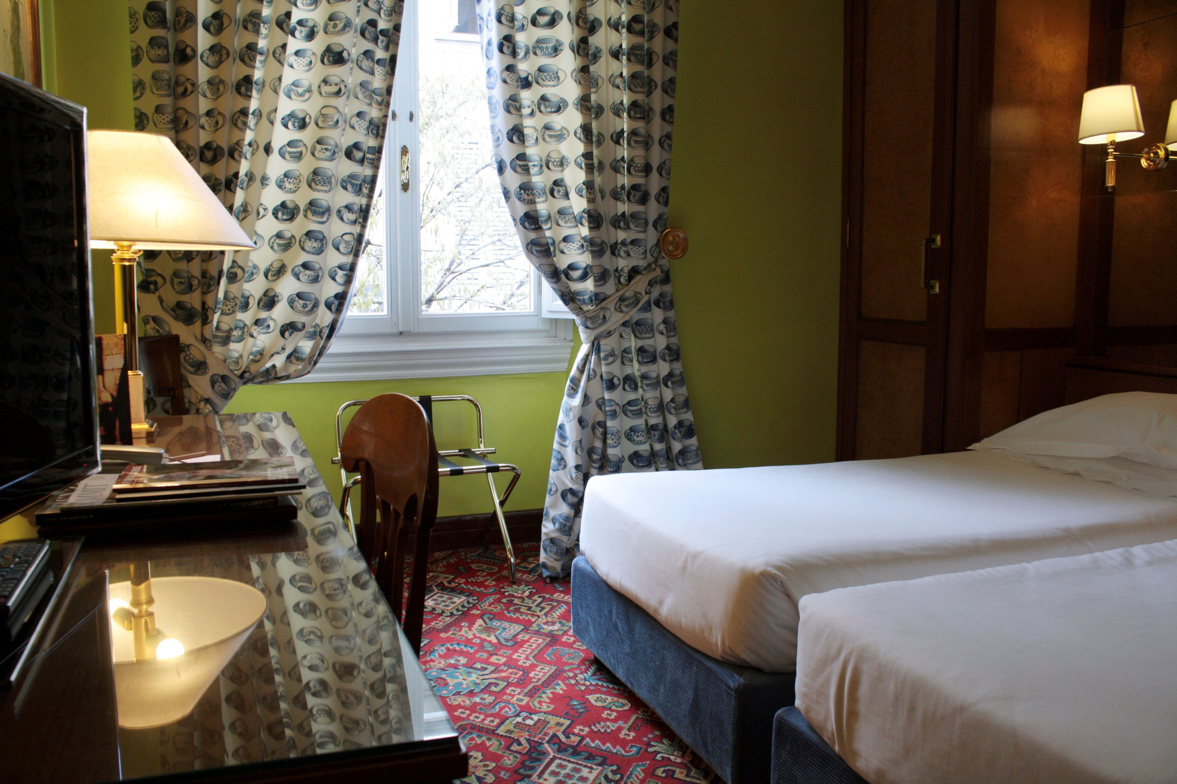 Een hotelkamer met een bed, een bureau met lamp, een stoel en een raam met behang gordijnen.