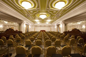 A conference room with rows of chairs and tables, a stage with a red carpet, and ornate chandeliers on the ceiling.