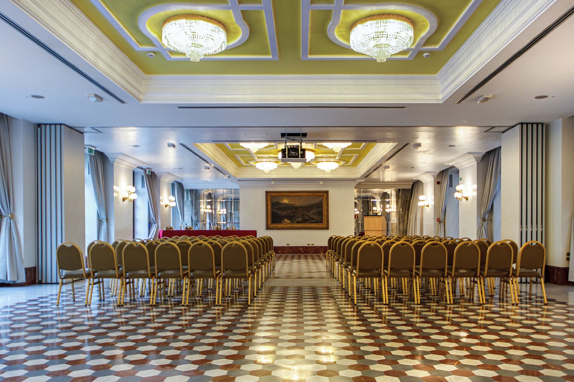 A conference room with rows of chairs, a patterned floor, chandeliers, and a painting on the wall.