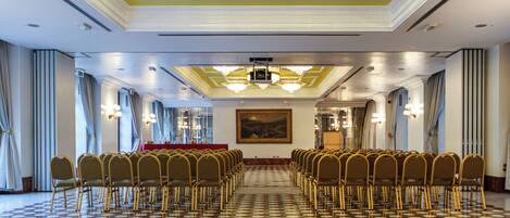 A conference room with rows of chairs, a patterned floor, chandeliers, and a painting on the wall.
