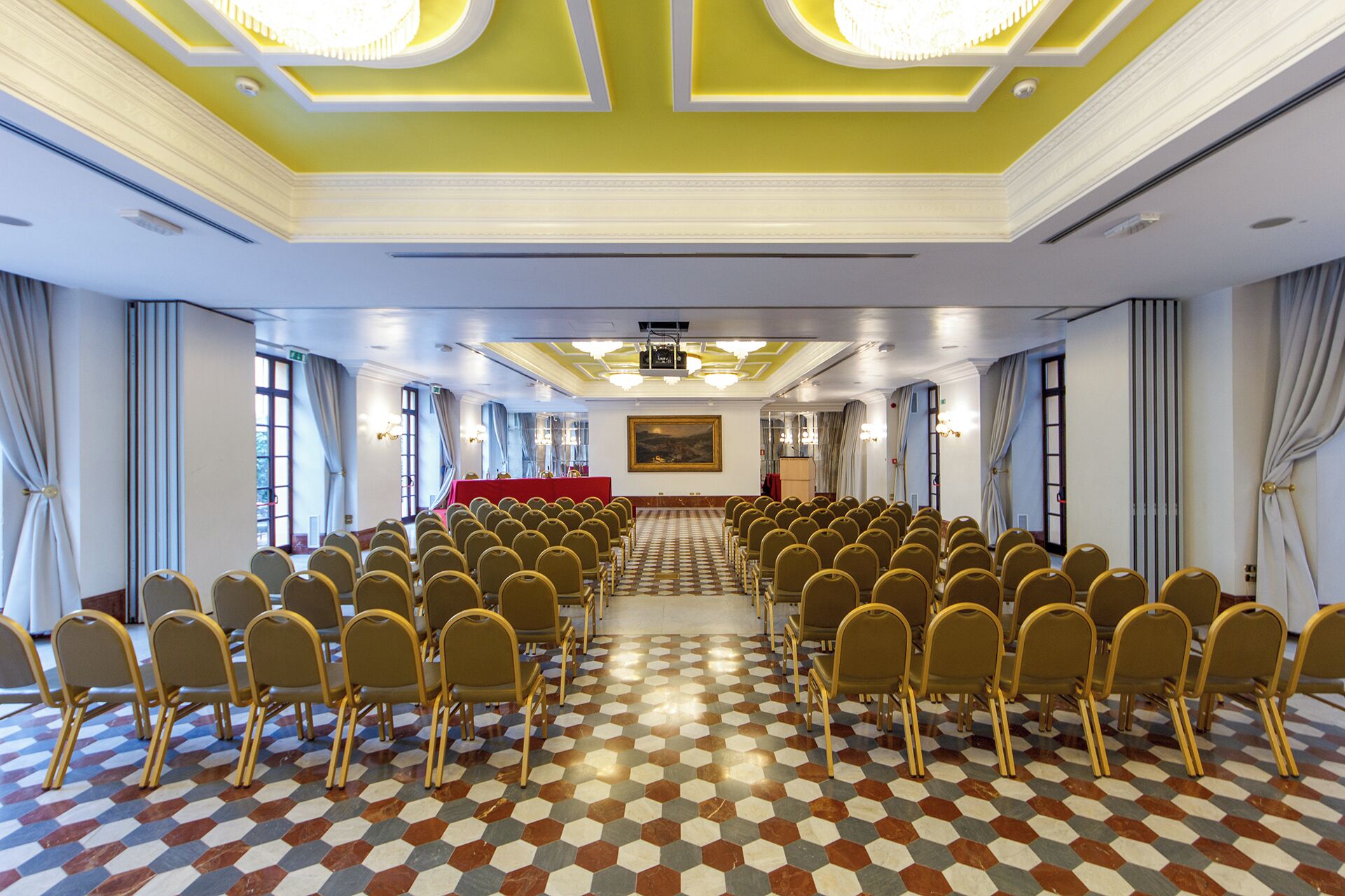 A conference room with rows of chairs, a stage with a red table, and a large painting on the wall.