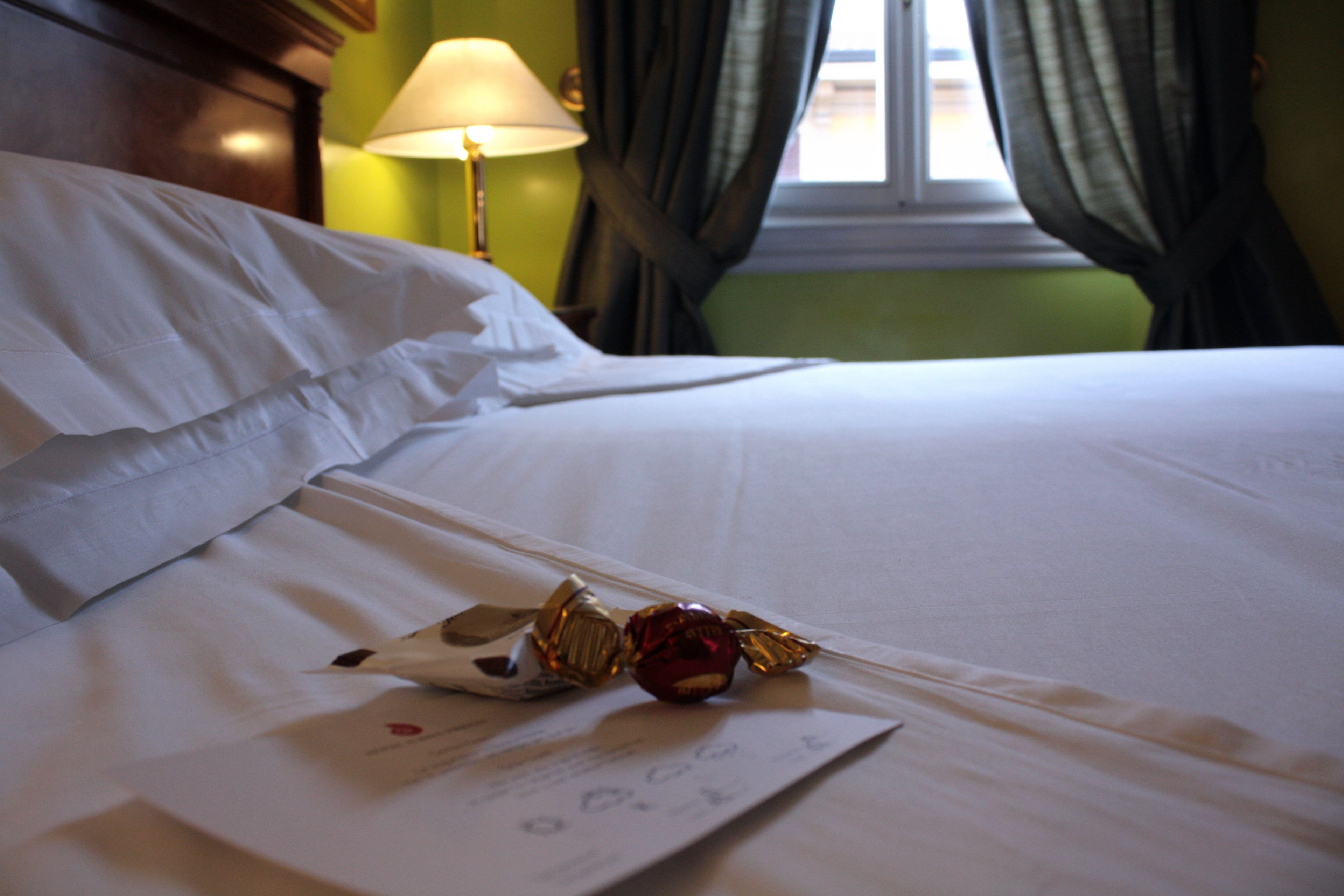 A neatly made bed with white linens, a lamp on the side, and a window with curtains.