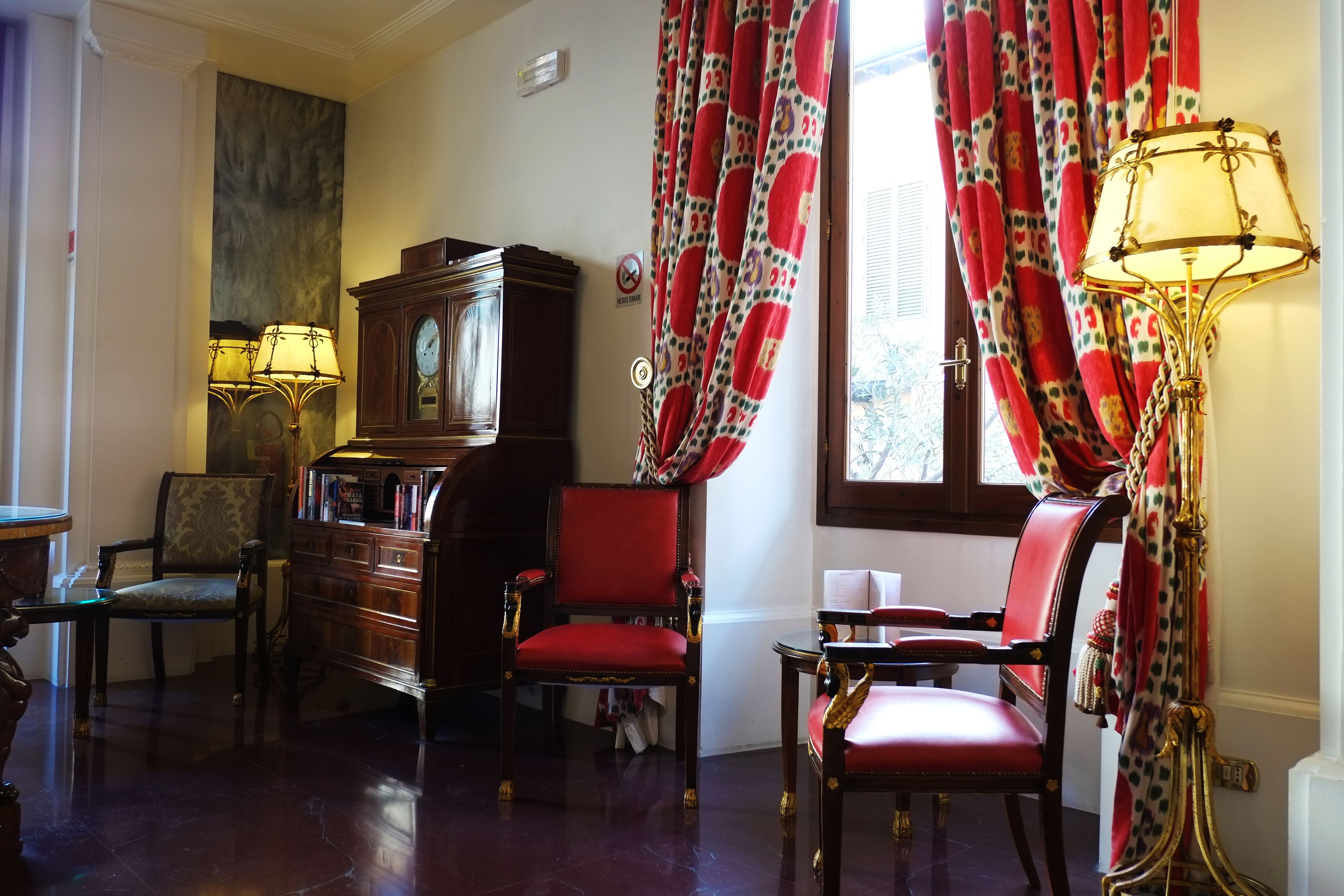 A room with a large wooden cabinet, two chairs, a red upholstered chair, and a table with a lamp.