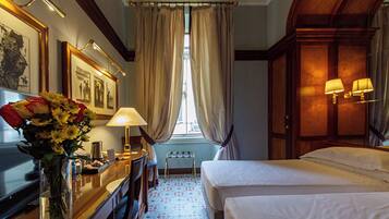 Une chambre d’hôtel avec deux lits, un bureau avec une lampe, un vase de fleurs et une fenêtre avec des rideaux.