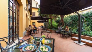 Un área de comedor al aire libre con mesas y sillas, rodeada de follaje y un piso de ladrillo.