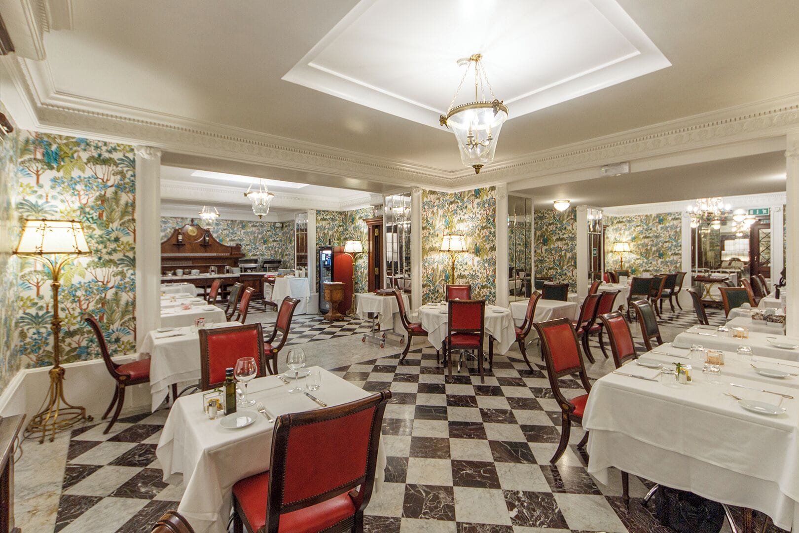 Une salle à manger élégante, avec un sol à carreaux, des nappes blanches et des chaises à l’assise rembourrée de tissu rouge.