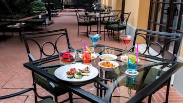 An outdoor dining area with glass tables and metal chairs, featuring a variety of drinks and snacks.