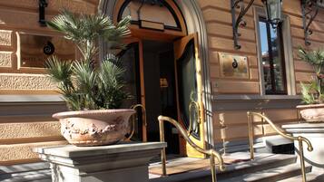 A hotel entrance with a large potted plant, a grand staircase with golden railings, and a plaque indicating the hotel name.