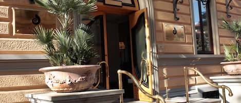 A hotel entrance with a large potted plant, a grand staircase with golden railings, and a plaque indicating the hotel name.