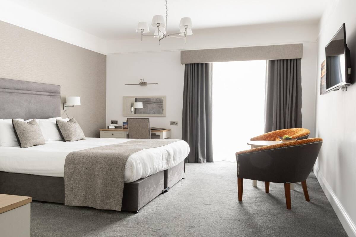 A modern hotel room with a large bed, a desk, a chair, a TV, and a window with curtains.