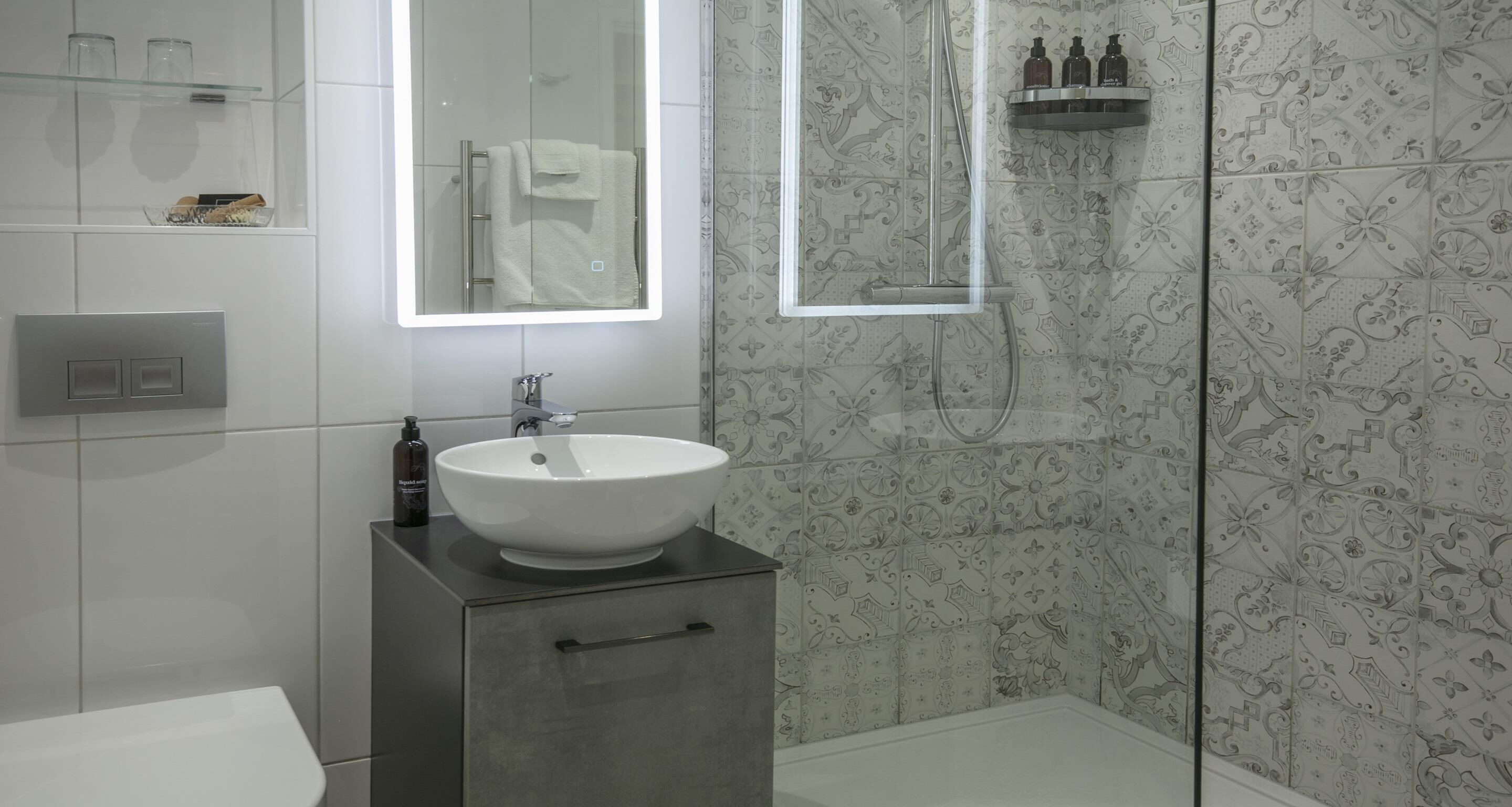 A bathroom with a white sink, a mirror above it, a shower with a glass door, and patterned tiles on the walls.