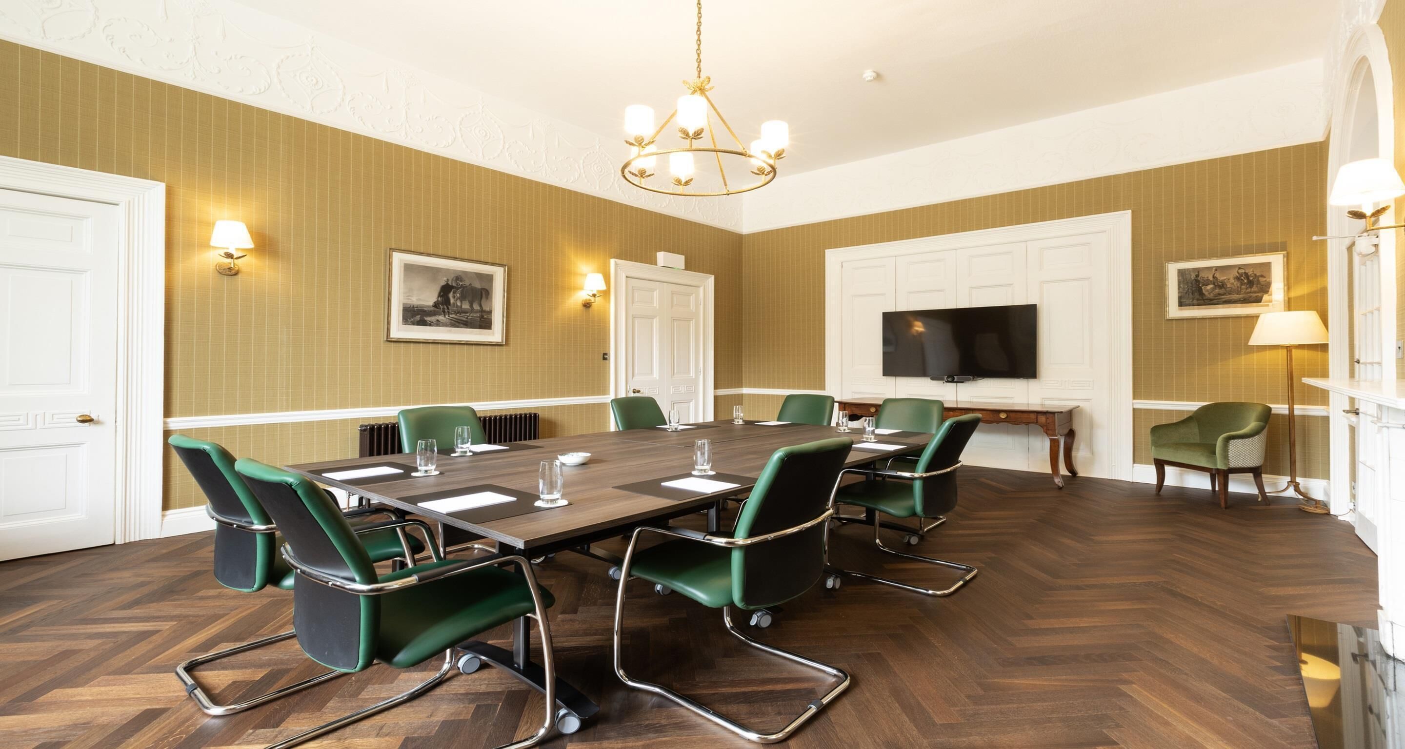A conference room with a large table, green chairs, a chandelier, and framed pictures on the walls.