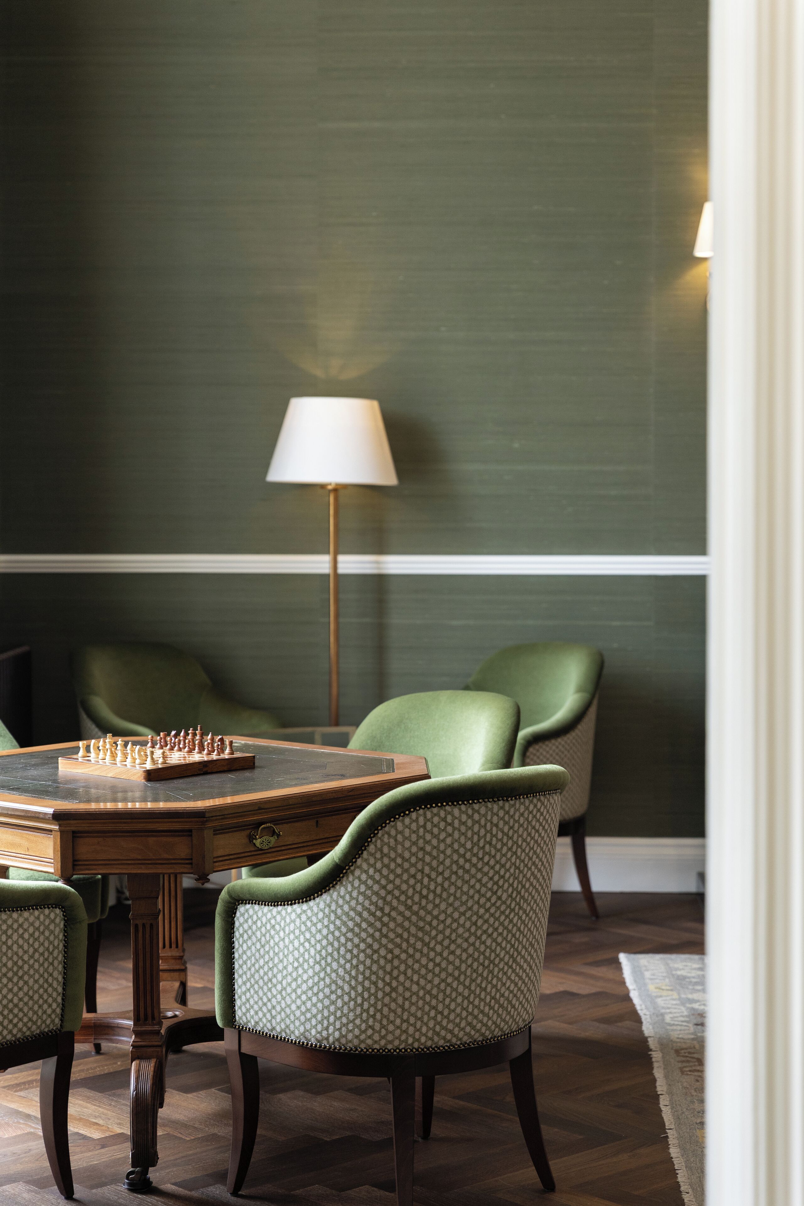 A dining area with a chessboard, a lamp, and green upholstered chairs.