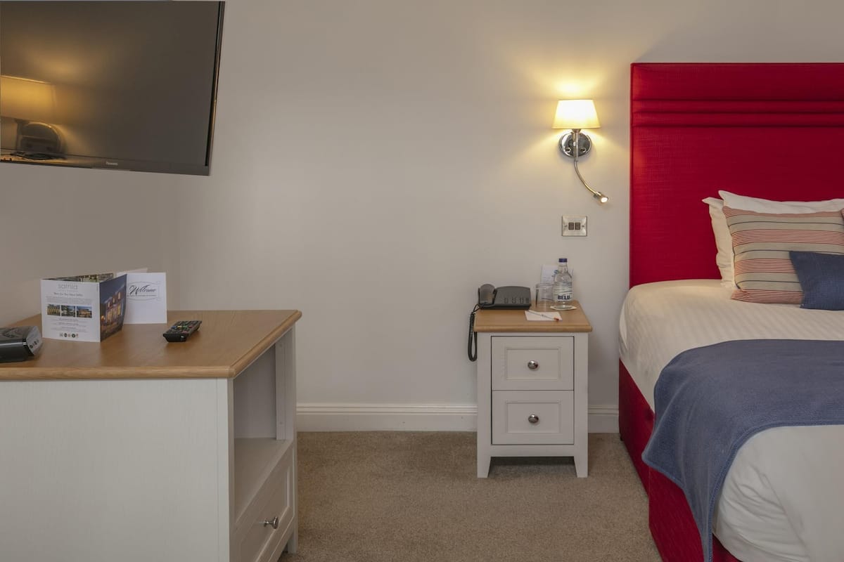 A hotel room with a bed, a nightstand, a lamp, a TV, and a desk with a remote control.