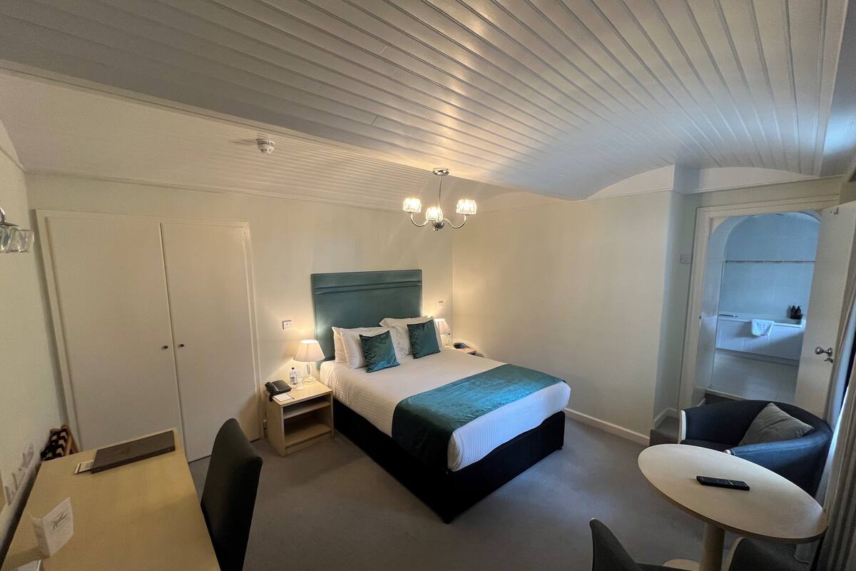 A hotel room with a bed, bedside table, lamp, chair, and a small round table.
