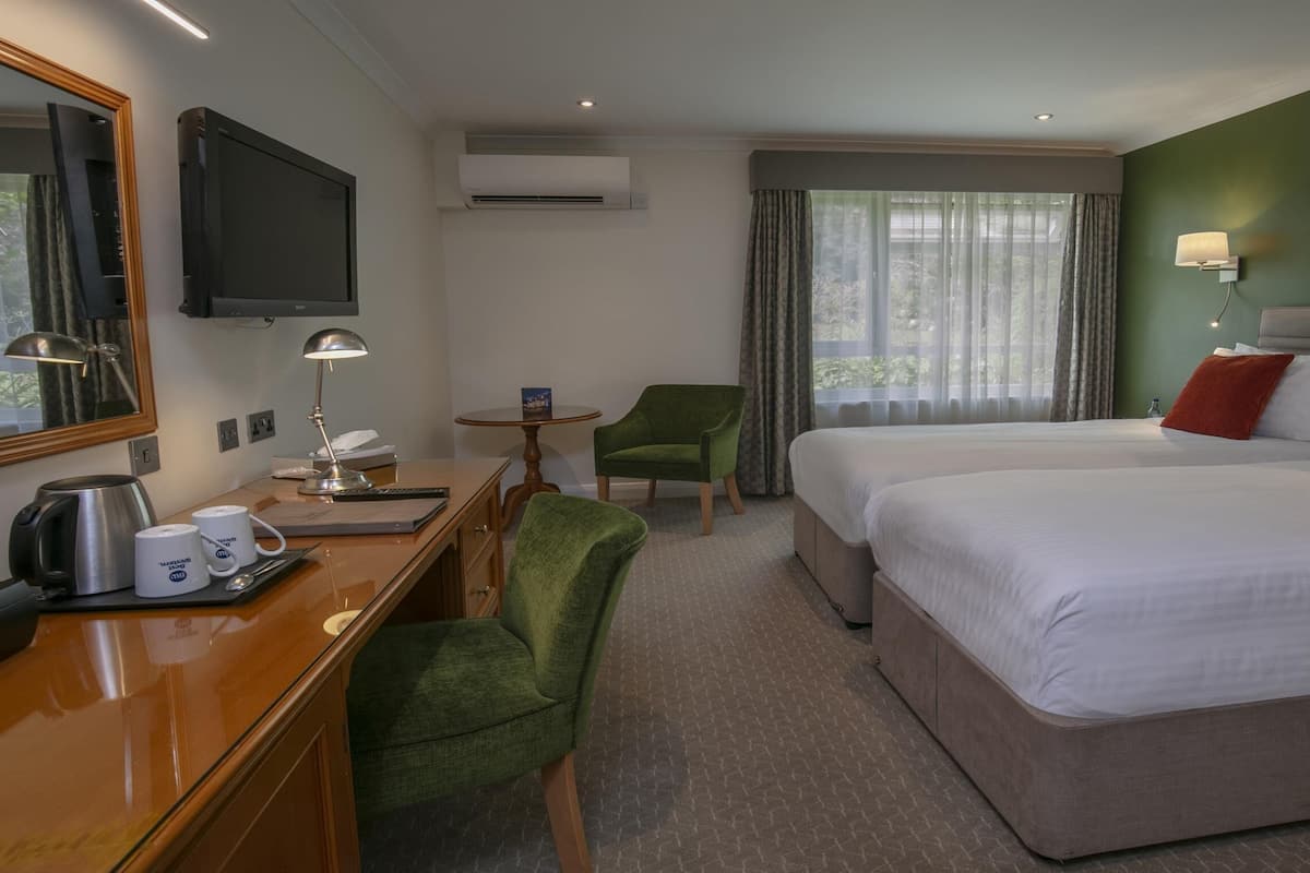 A hotel room with a bed, two green chairs, a desk with a coffee maker, a TV, and a window with curtains.