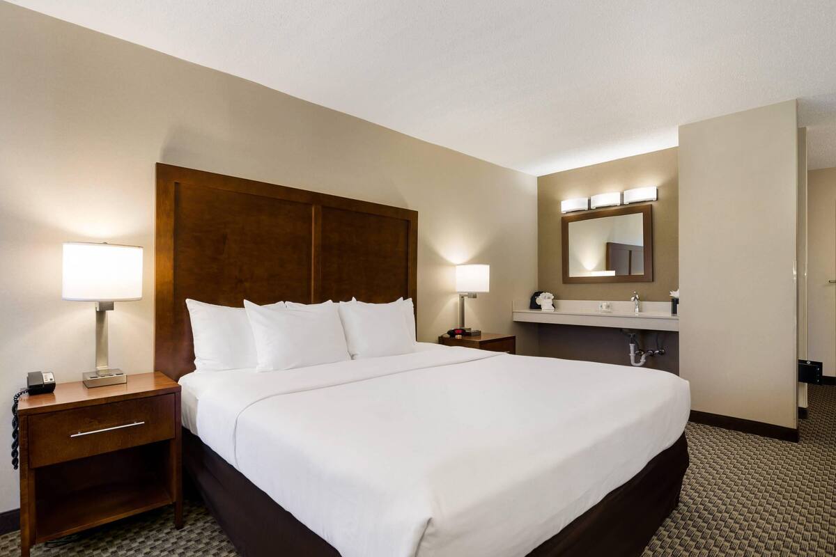 A hotel room with a large bed, a wooden headboard, a nightstand with a lamp, a bathroom with a mirror and lights, and a carpeted floor.