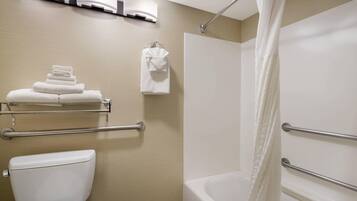 A bathroom with a toilet, a bathtub with a shower curtain, a towel rack with folded towels, and a handrail.
