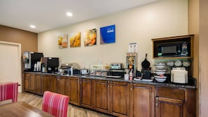 A hotel kitchen with wooden cabinets, a microwave, a coffee machine, and a toaster. There are framed pictures on the wall and a dining area with a table and chairs. - Comfort Inn Lexington Virginia Horse Center (Lexington)