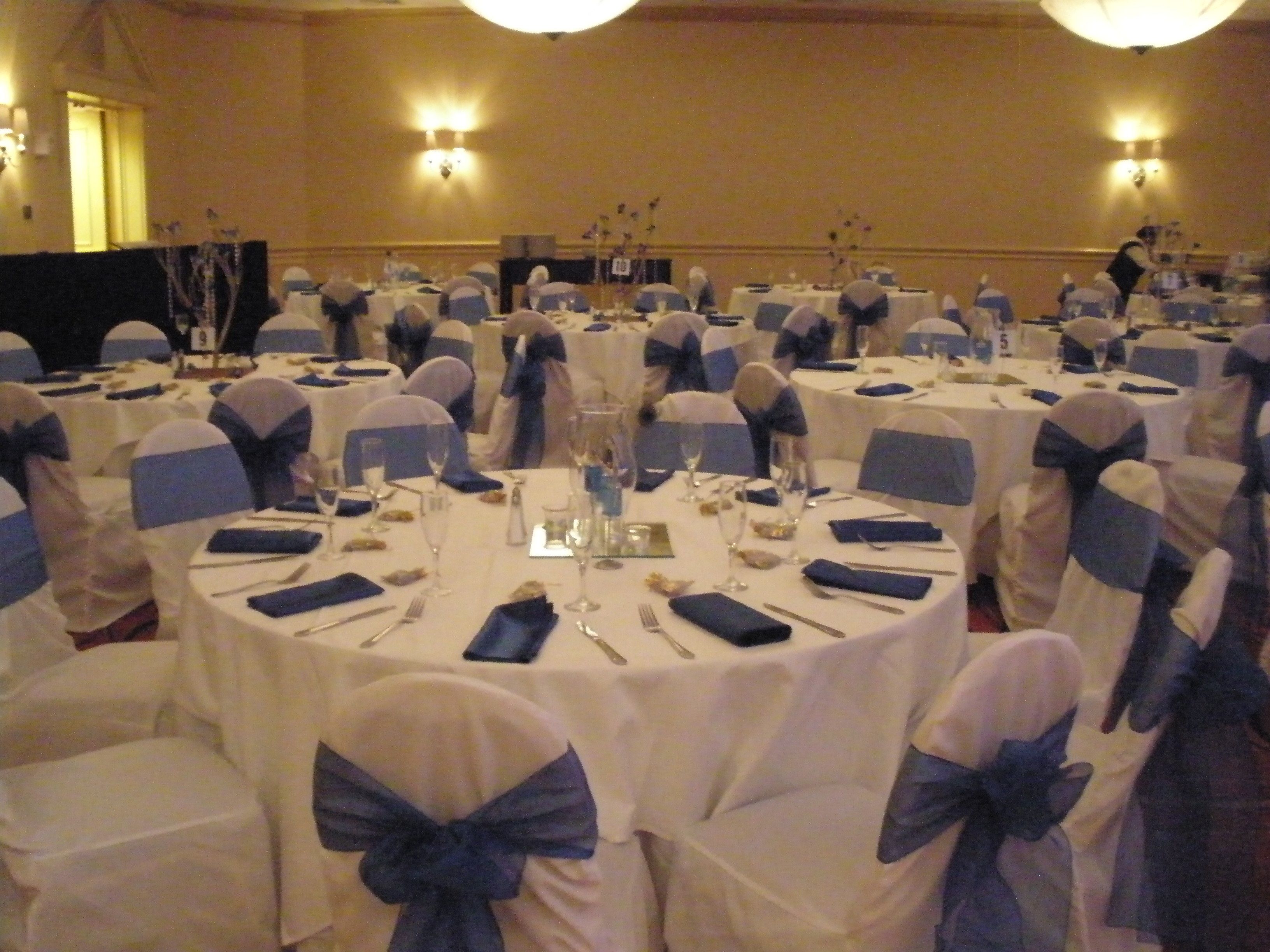 A banquet hall with round tables set for a formal event, each with white tablecloths and blue sashes on the chairs.