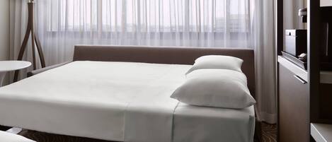 A hotel room with a bed, a bedside table, a lamp, and a view through sheer curtains.