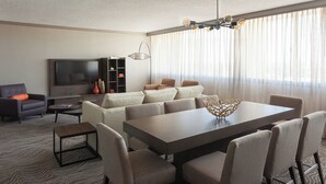 A modern living room with a large flat-screen TV, a sofa set, a dining table with chairs, and a patterned carpet.
