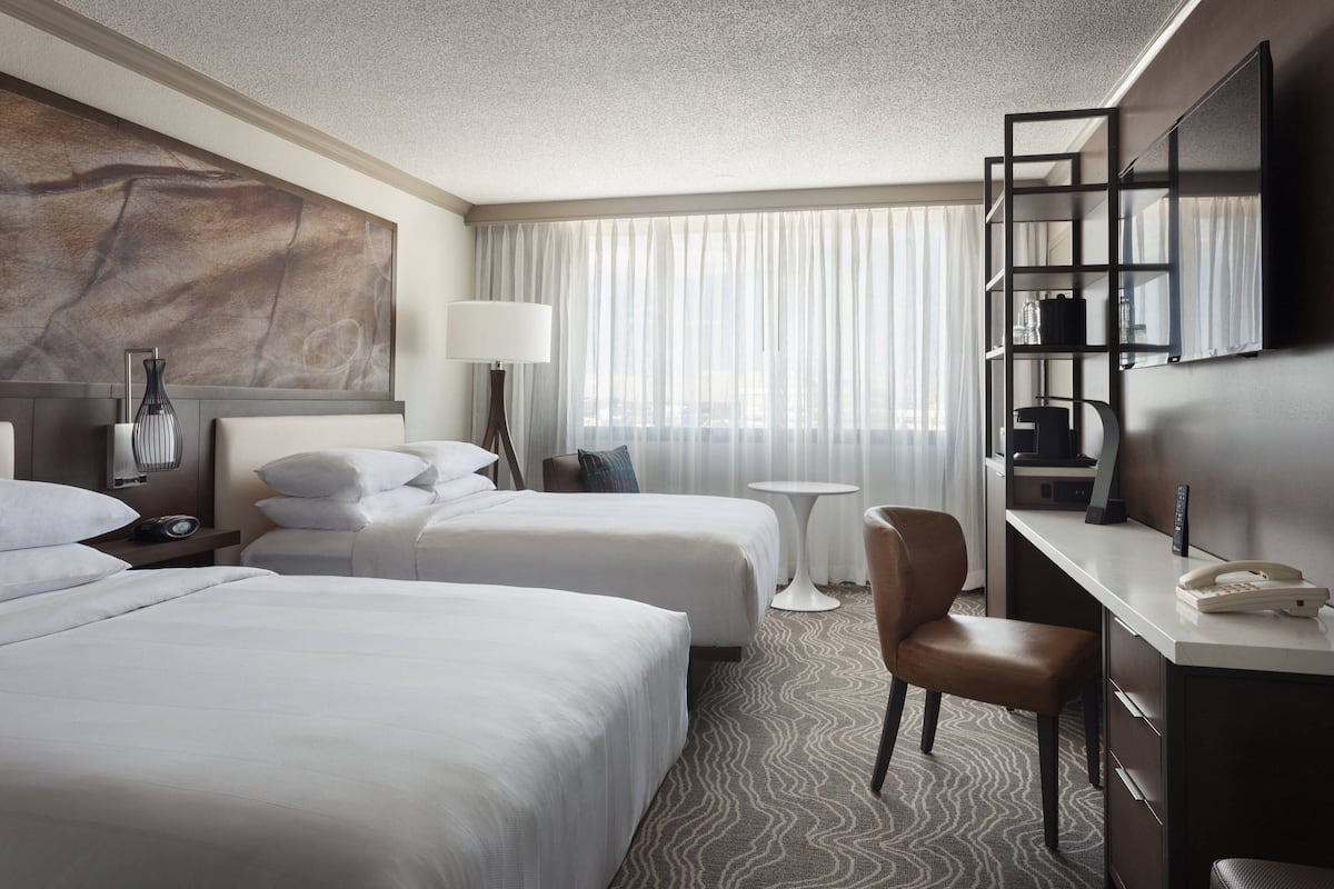 A hotel room with two beds, a desk, a chair, a small table, a lamp, a telephone, and a large abstract painting on the wall.