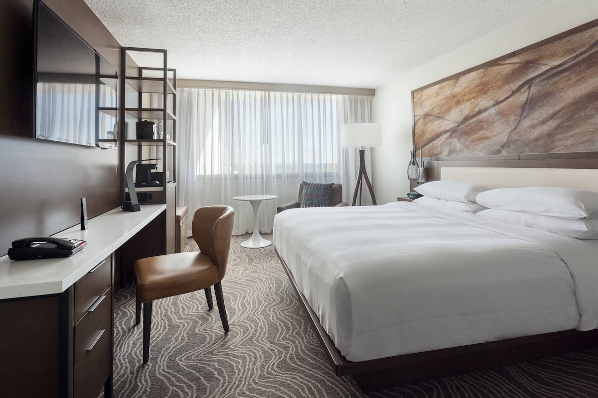 A hotel room with a bed, a desk, a chair, a small table, and a TV mounted on the wall.