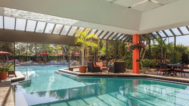 A swimming pool with clear water, surrounded by lounge chairs and umbrellas, and a glass ceiling above.