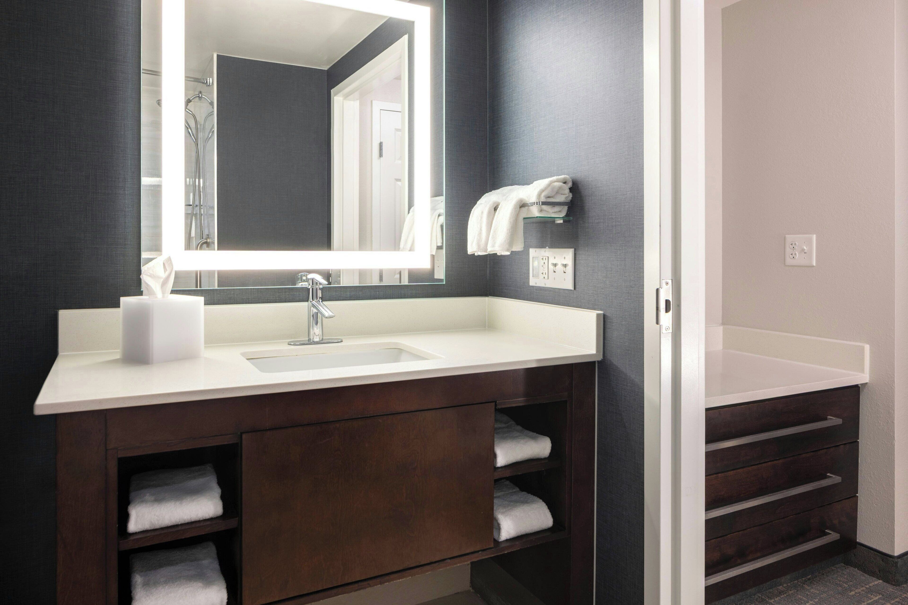 A modern bathroom with a large mirror, white sink, dark wooden vanity, and built-in shelves for towels.