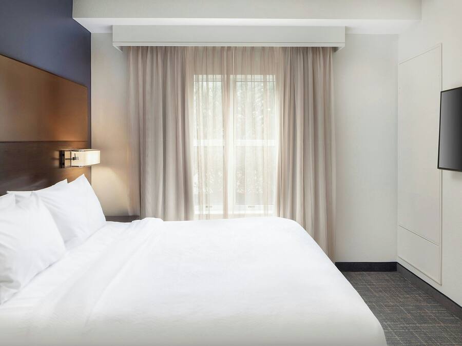 A hotel room with a large bed, a flat-screen TV mounted on the wall, and a window with sheer curtains.