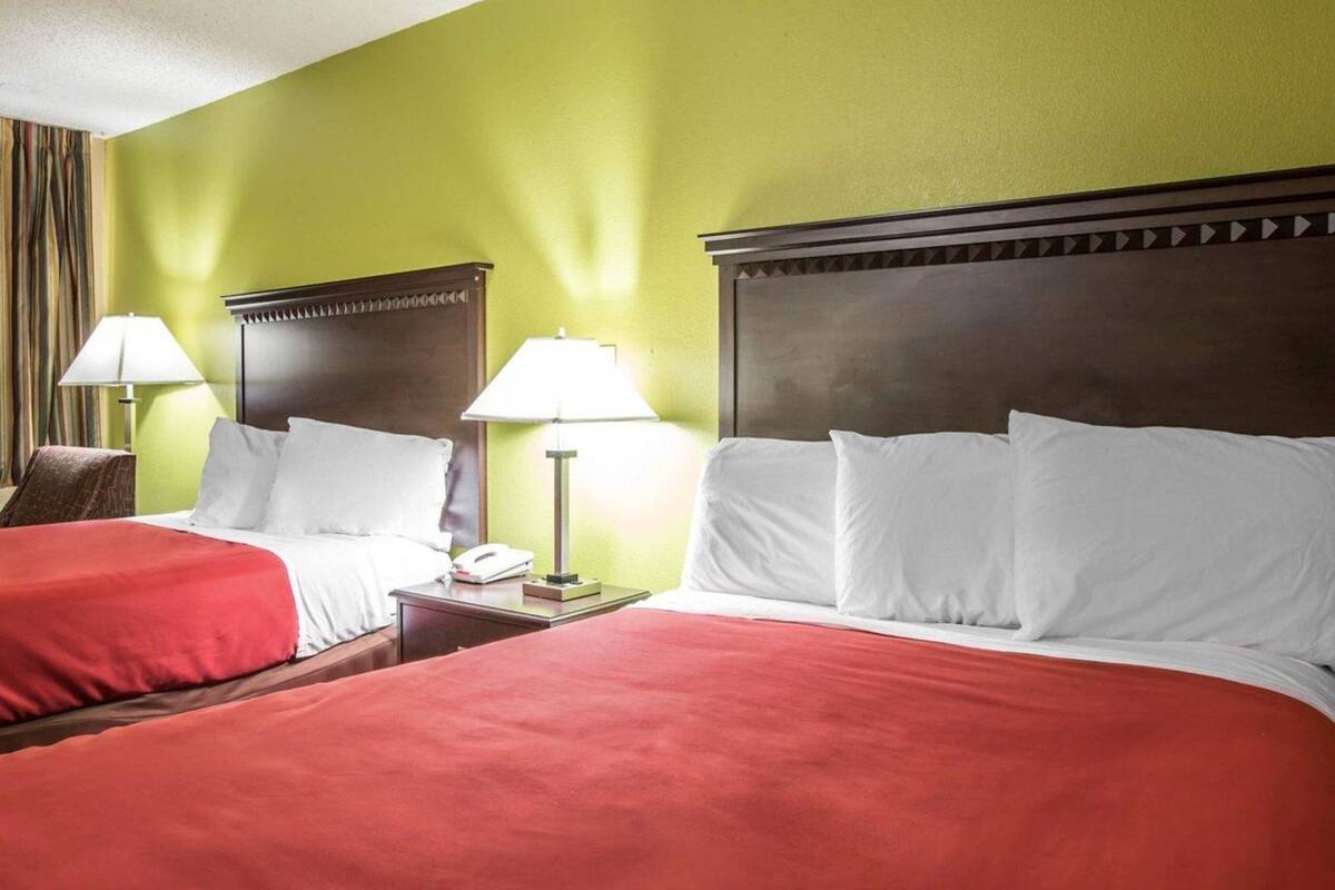 A hotel room with two beds, each with red bedding, a wooden headboard, and a nightstand with a lamp.